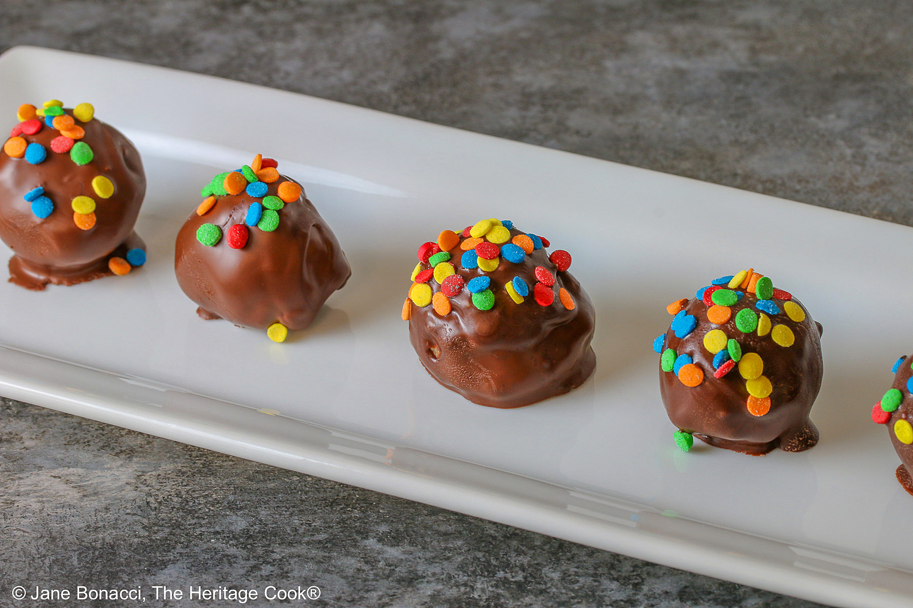 A line of five Chocolate Chip Cookie Dough Truffles on a white rectangular platter, each one dipped in chocolate with brightly colored confetti candy sprinkles on top of each © 2026 Jane Bonacci, The Heritage Cook.