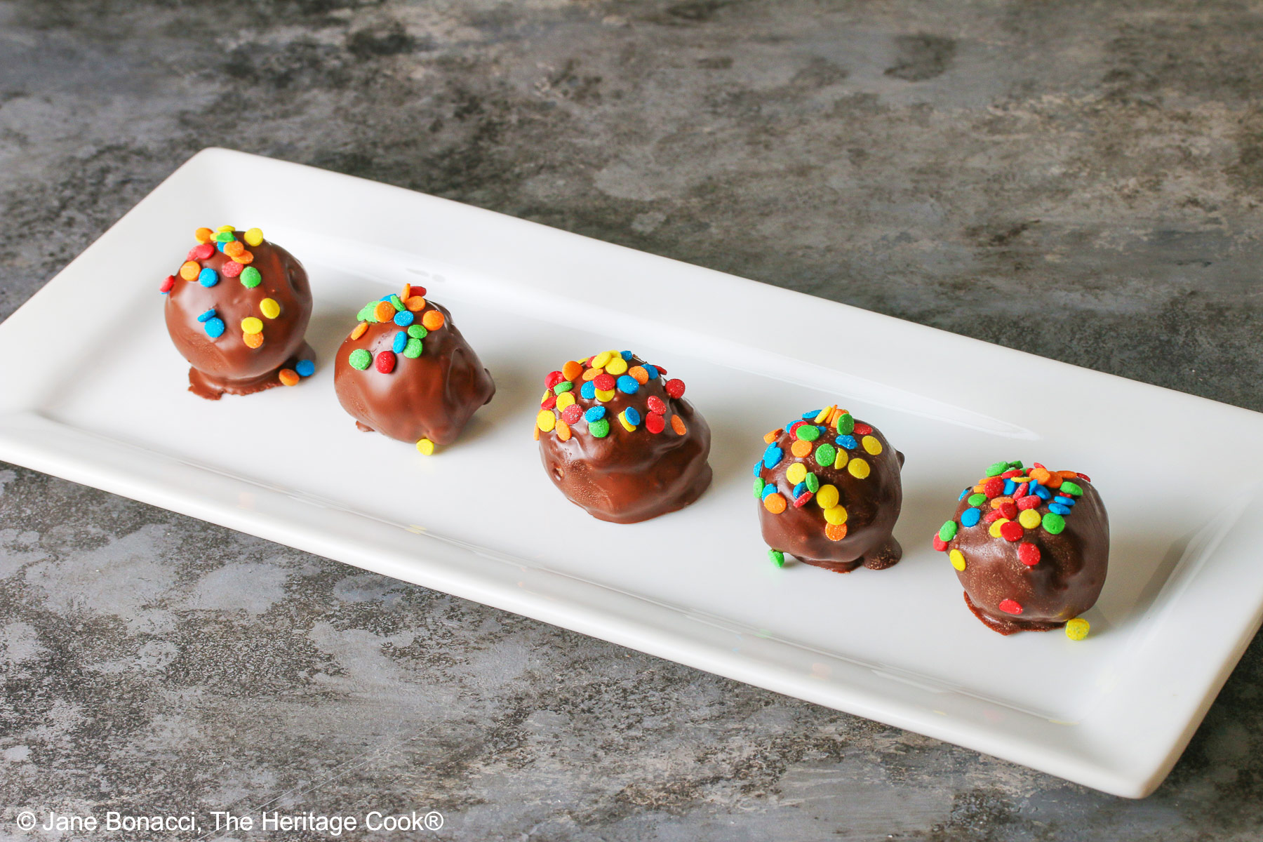 A line of five Chocolate Chip Cookie Dough Truffles on a white rectangular platter, each one dipped in chocolate with brightly colored confetti candy sprinkles on top of each © 2026 Jane Bonacci, The Heritage Cook.