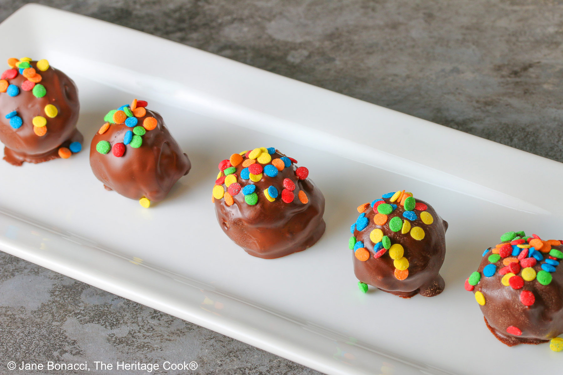 A line of five Chocolate Chip Cookie Dough Truffles on a white rectangular platter, each one dipped in chocolate with brightly colored confetti candy sprinkles on top of each © 2026 Jane Bonacci, The Heritage Cook.