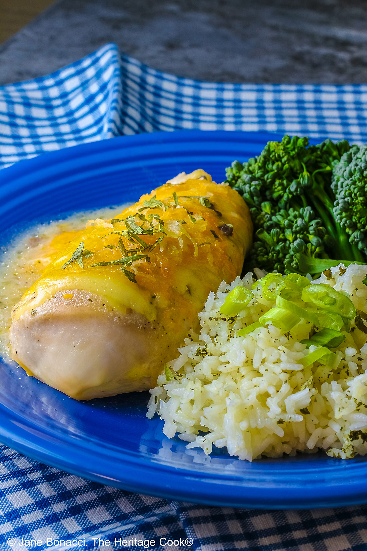 Large boneless chicken breast on a dark blue plate, topped with sauce and melted cheese, sprinkled with tarragon leaves, served with green broccolini and steamed rice dotted with herbs and sliced green onion on top, sitting on a blue and white checked cloth © 2026 Jane Bonacci, The Heritage Cook.