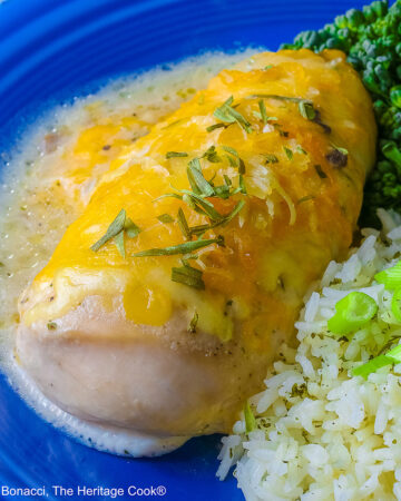 Large boneless chicken breast on a dark blue plate, topped with sauce and melted cheese, sprinkled with tarragon leaves, served with green broccolini and steamed rice dotted with herbs and sliced green onion on top, sitting on a blue and white checked cloth © 2026 Jane Bonacci, The Heritage Cook.
