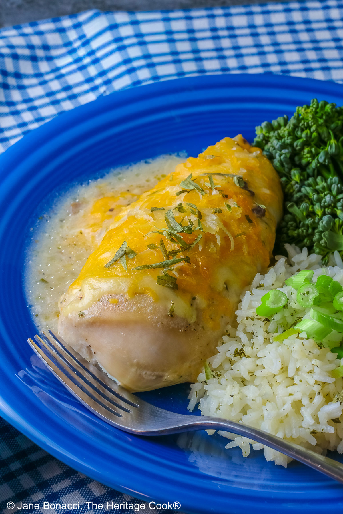 Large boneless chicken breast on a dark blue plate, topped with sauce and melted cheese, sprinkled with tarragon leaves, served with green broccolini and steamed rice dotted with herbs and sliced green onion on top, sitting on a blue and white checked cloth © 2026 Jane Bonacci, The Heritage Cook.