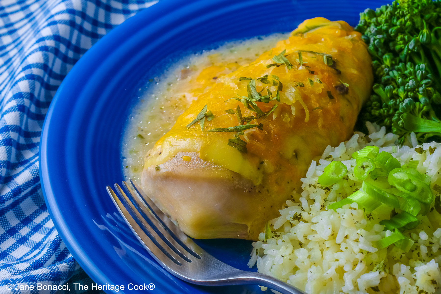 Large boneless chicken breast on a dark blue plate, topped with sauce and melted cheese, sprinkled with tarragon leaves, served with green broccolini and steamed rice dotted with herbs and sliced green onion on top, sitting on a blue and white checked cloth © 2026 Jane Bonacci, The Heritage Cook.