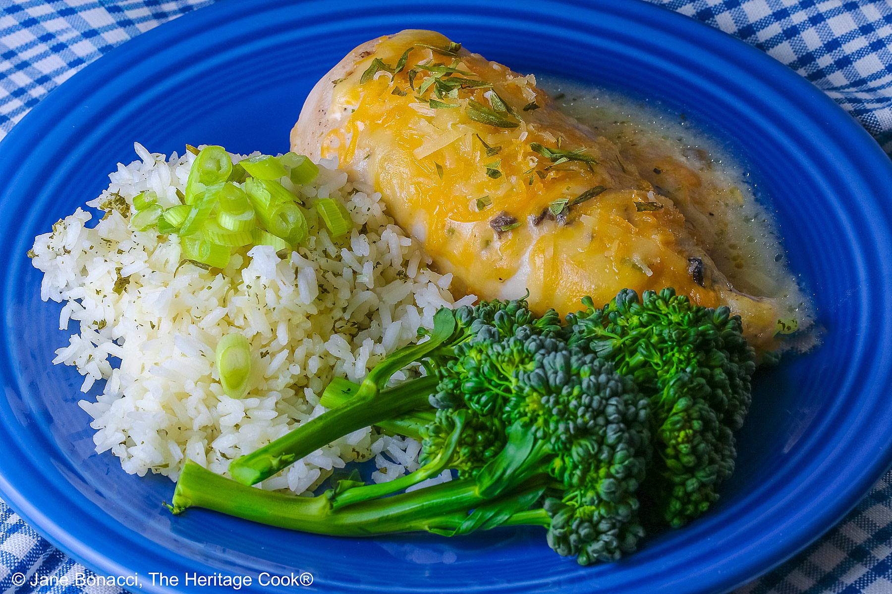 Large boneless chicken breast on a dark blue plate, topped with sauce and melted cheese, sprinkled with tarragon leaves, served with green broccolini and steamed rice dotted with herbs and sliced green onion on top, sitting on a blue and white checked cloth © 2026 Jane Bonacci, The Heritage Cook.