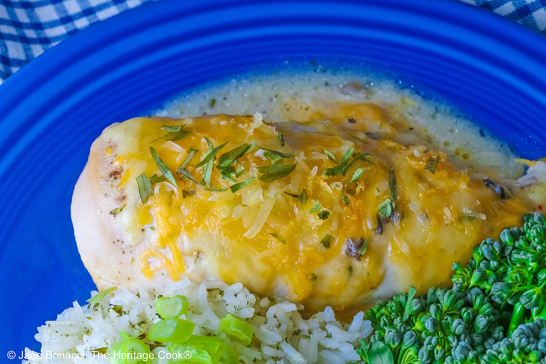 Large boneless chicken breast on a dark blue plate, topped with sauce and melted cheese, sprinkled with tarragon leaves, served with green broccolini and steamed rice dotted with herbs and sliced green onion on top, sitting on a blue and white checked cloth © 2026 Jane Bonacci, The Heritage Cook.