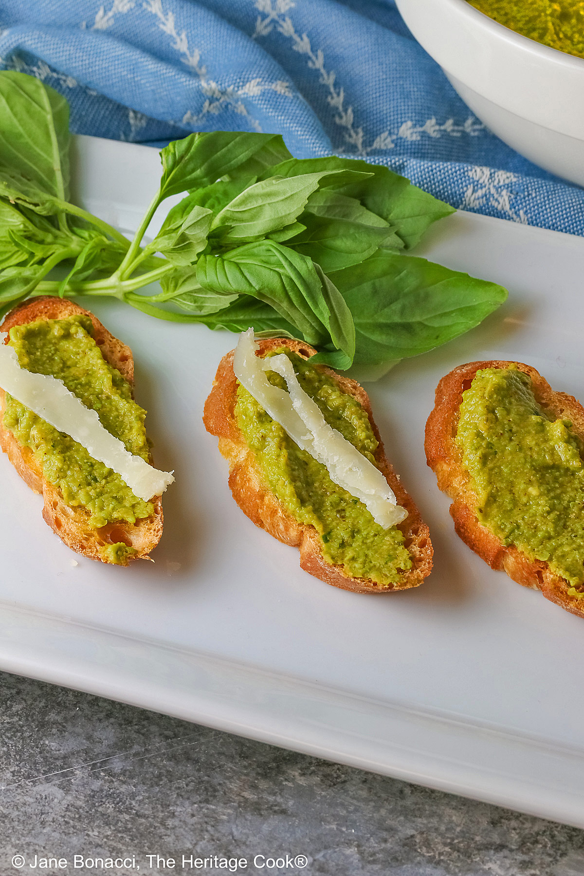A large bowl of Mediterranean Pea Puree sitting on a blue cloth behind a rectangular white platter with 5 of the crostini, each topped with a shaving of Parmesan cheese and some fresh basil on the side © 2026 Jane Bonacci, The Heritage Cook.