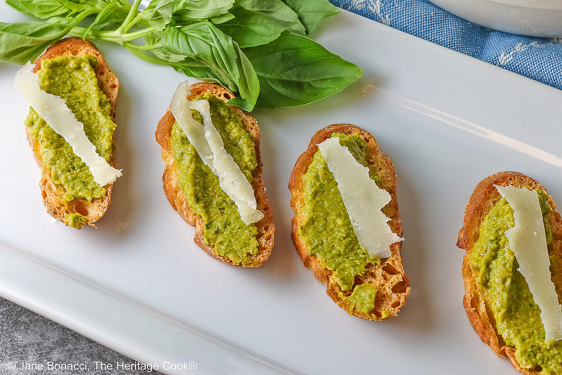 A large bowl of Mediterranean Pea Puree sitting on a blue cloth behind a rectangular white platter with 5 of the crostini, each topped with a shaving of Parmesan cheese and some fresh basil on the side © 2026 Jane Bonacci, The Heritage Cook.