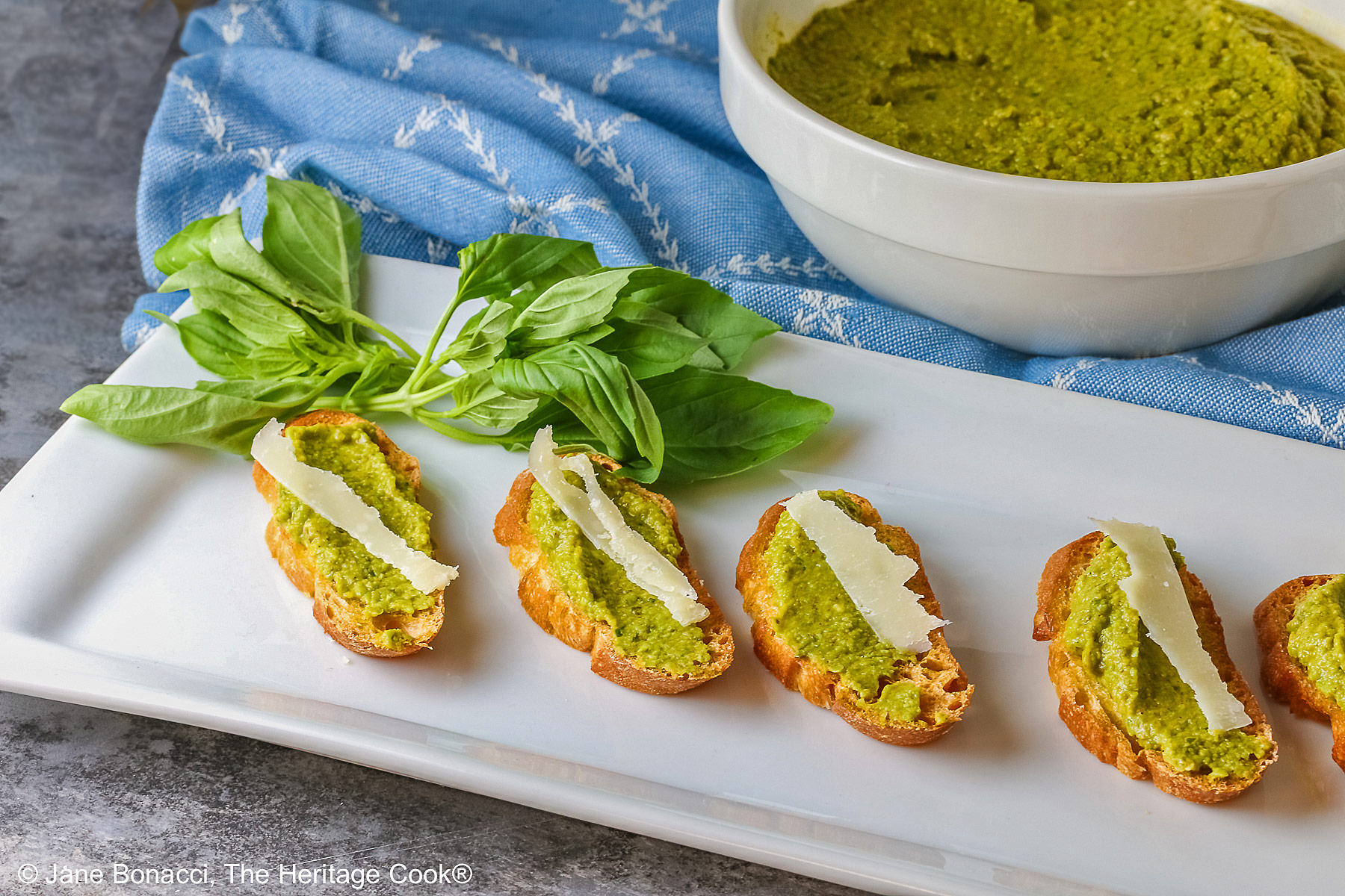 A large bowl of Mediterranean Pea Puree sitting on a blue cloth behind a rectangular white platter with 5 of the crostini, each topped with a shaving of Parmesan cheese and some fresh basil on the side © 2026 Jane Bonacci, The Heritage Cook.