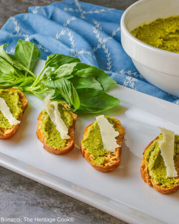 A large bowl of Mediterranean Pea Puree sitting on a blue cloth behind a rectangular white platter with 5 of the crostini, each topped with a shaving of Parmesan cheese and some fresh basil on the side © 2026 Jane Bonacci, The Heritage Cook.
