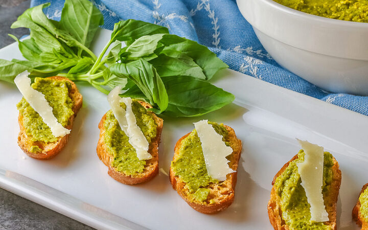 A large bowl of Mediterranean Pea Puree sitting on a blue cloth behind a rectangular white platter with 5 of the crostini, each topped with a shaving of Parmesan cheese and some fresh basil on the side © 2026 Jane Bonacci, The Heritage Cook.