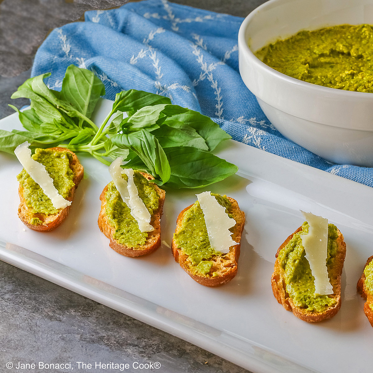 A large bowl of Mediterranean Pea Puree sitting on a blue cloth behind a rectangular white platter with 5 of the crostini, each topped with a shaving of Parmesan cheese and some fresh basil on the side © 2026 Jane Bonacci, The Heritage Cook.