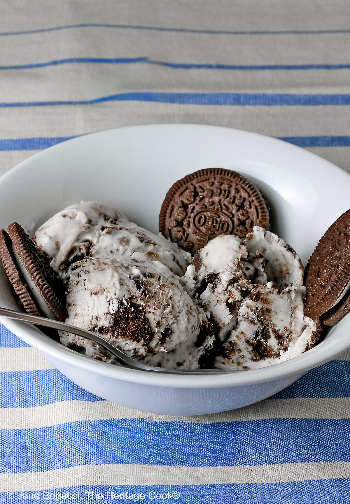 White bowl filled with 3 scoops of No Churn Cookies and Cream Cheesecake Ice Cream with several whole cookies tucked into the bowl and a spoon resting alongside one of the scoops, sitting on a blue and white striped cloth © 2026 Jane Bonacci, The Heritage Cook.