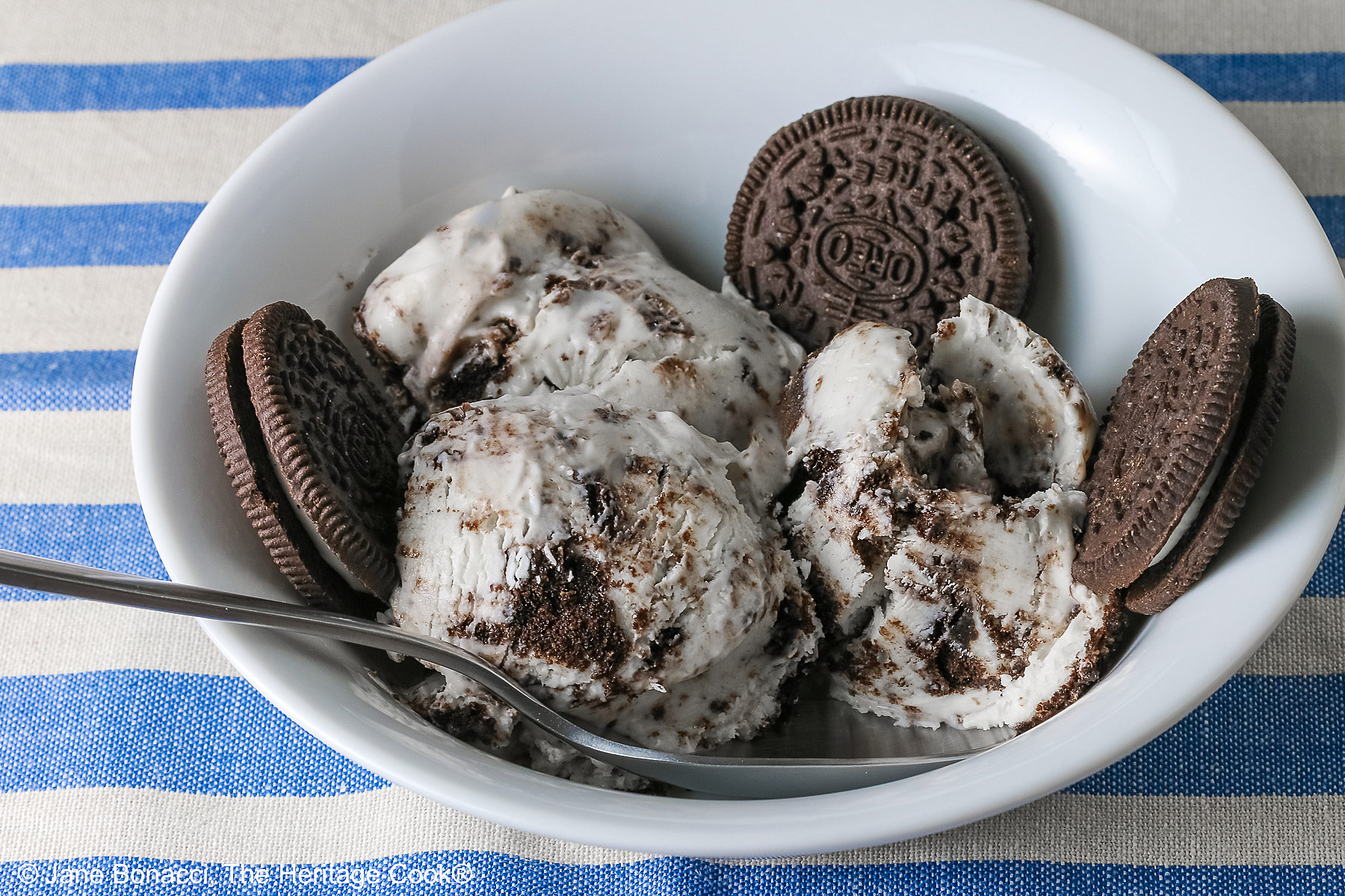 White bowl filled with 3 scoops of No Churn Cookies and Cream Cheesecake Ice Cream with several whole cookies tucked into the bowl and a spoon resting alongside one of the scoops, sitting on a blue and white striped cloth © 2026 Jane Bonacci, The Heritage Cook.