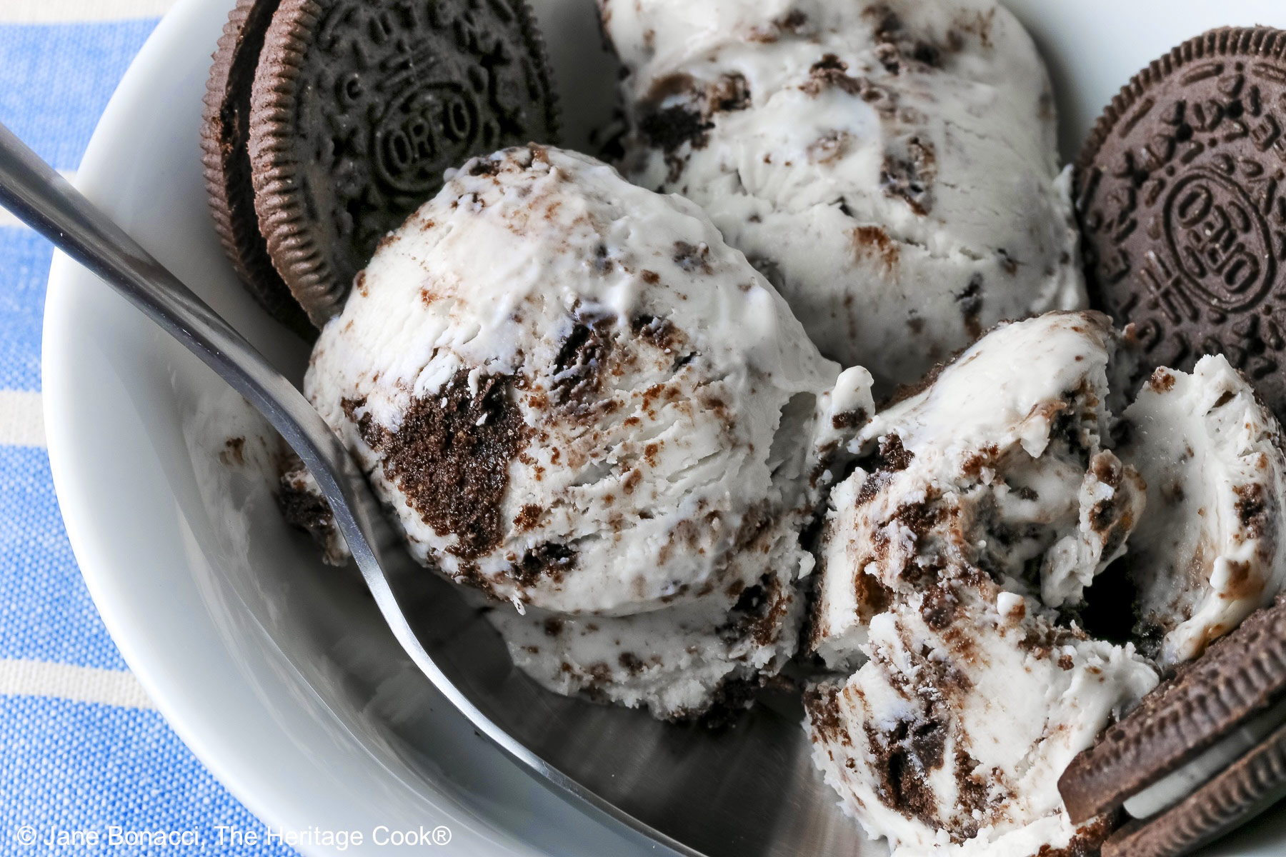 White bowl filled with 3 scoops of No Churn Cookies and Cream Cheesecake Ice Cream with several whole cookies tucked into the bowl and a spoon resting alongside one of the scoops, sitting on a blue and white striped cloth © 2026 Jane Bonacci, The Heritage Cook.