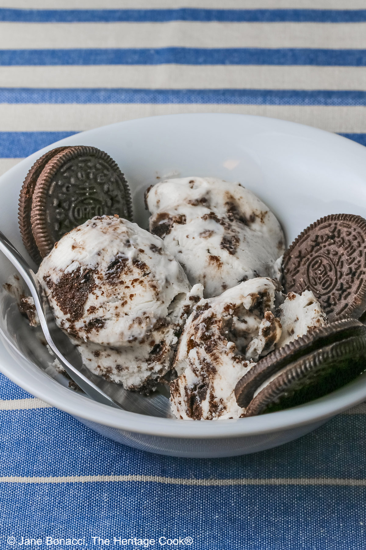 White bowl filled with 3 scoops of No Churn Cookies and Cream Cheesecake Ice Cream with several whole cookies tucked into the bowl and a spoon resting alongside one of the scoops, sitting on a blue and white striped cloth © 2026 Jane Bonacci, The Heritage Cook.