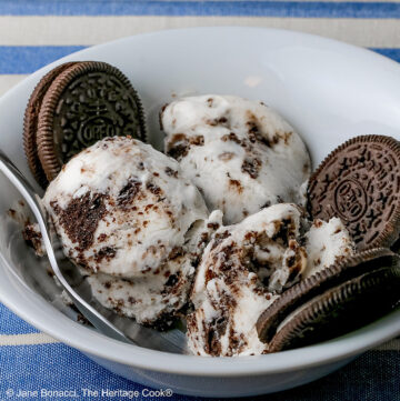 White bowl filled with 3 scoops of No Churn Cookies and Cream Cheesecake Ice Cream with several whole cookies tucked into the bowl and a spoon resting alongside one of the scoops, sitting on a blue and white striped cloth © 2026 Jane Bonacci, The Heritage Cook.