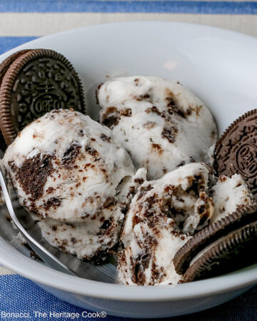 White bowl filled with 3 scoops of No Churn Cookies and Cream Cheesecake Ice Cream with several whole cookies tucked into the bowl and a spoon resting alongside one of the scoops, sitting on a blue and white striped cloth © 2026 Jane Bonacci, The Heritage Cook.