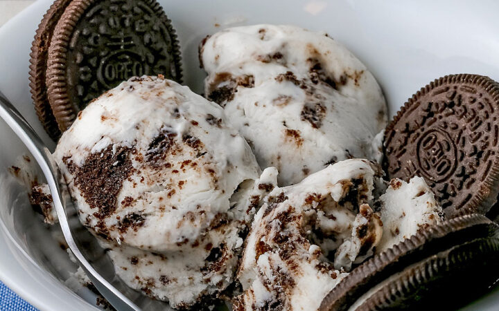 White bowl filled with 3 scoops of No Churn Cookies and Cream Cheesecake Ice Cream with several whole cookies tucked into the bowl and a spoon resting alongside one of the scoops, sitting on a blue and white striped cloth © 2026 Jane Bonacci, The Heritage Cook.