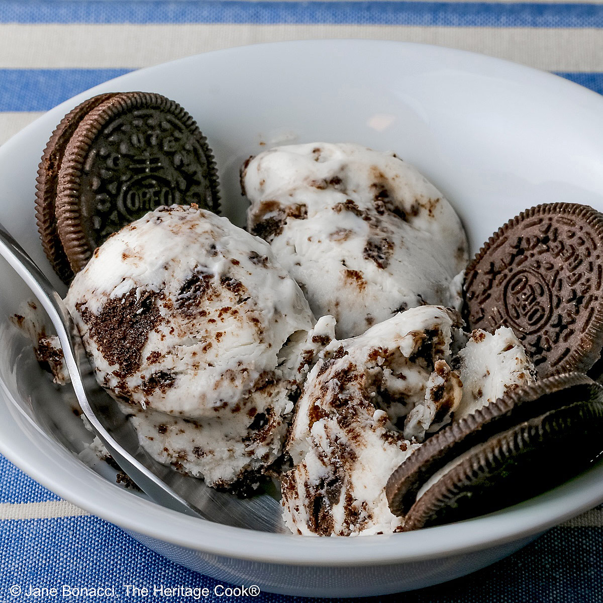 White bowl filled with 3 scoops of No Churn Cookies and Cream Cheesecake Ice Cream with several whole cookies tucked into the bowl and a spoon resting alongside one of the scoops, sitting on a blue and white striped cloth © 2026 Jane Bonacci, The Heritage Cook.