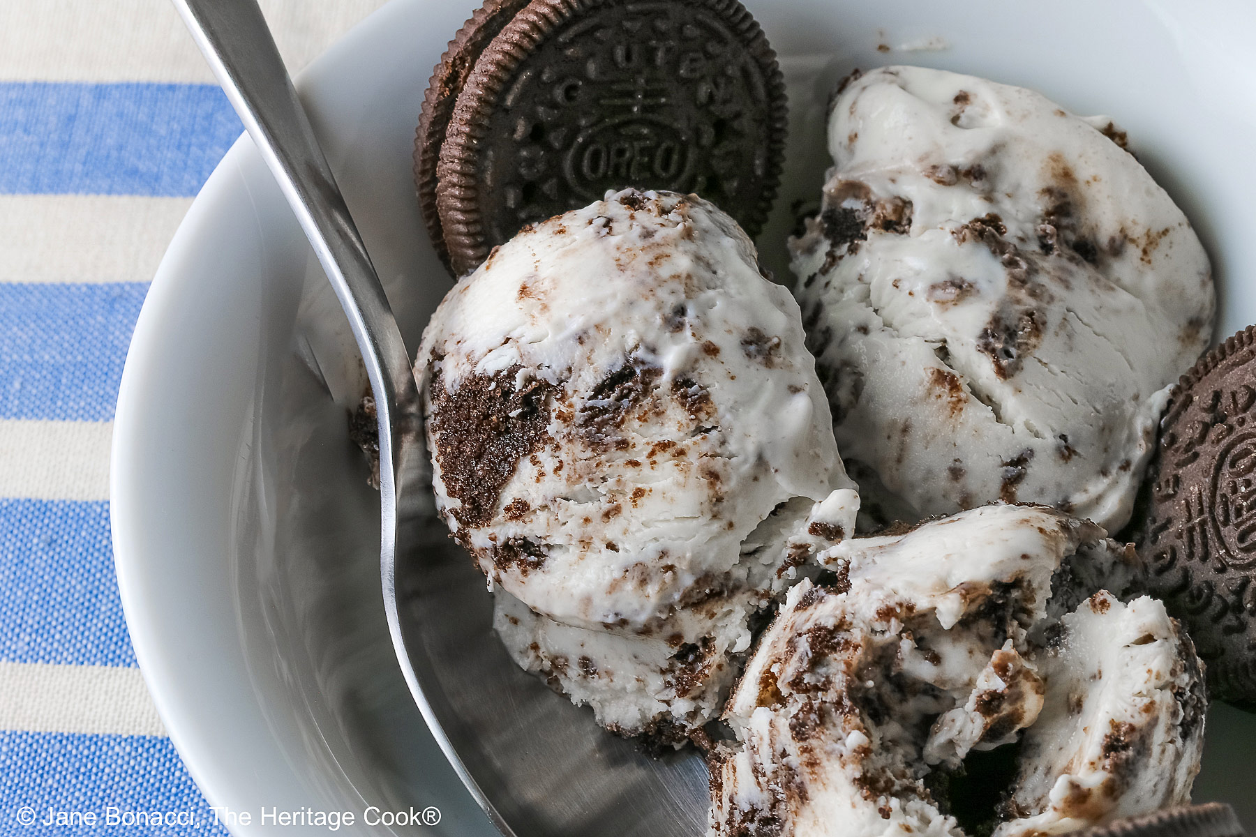 White bowl filled with 3 scoops of No Churn Cookies and Cream Cheesecake Ice Cream with several whole cookies tucked into the bowl and a spoon resting alongside one of the scoops, sitting on a blue and white striped cloth © 2026 Jane Bonacci, The Heritage Cook.