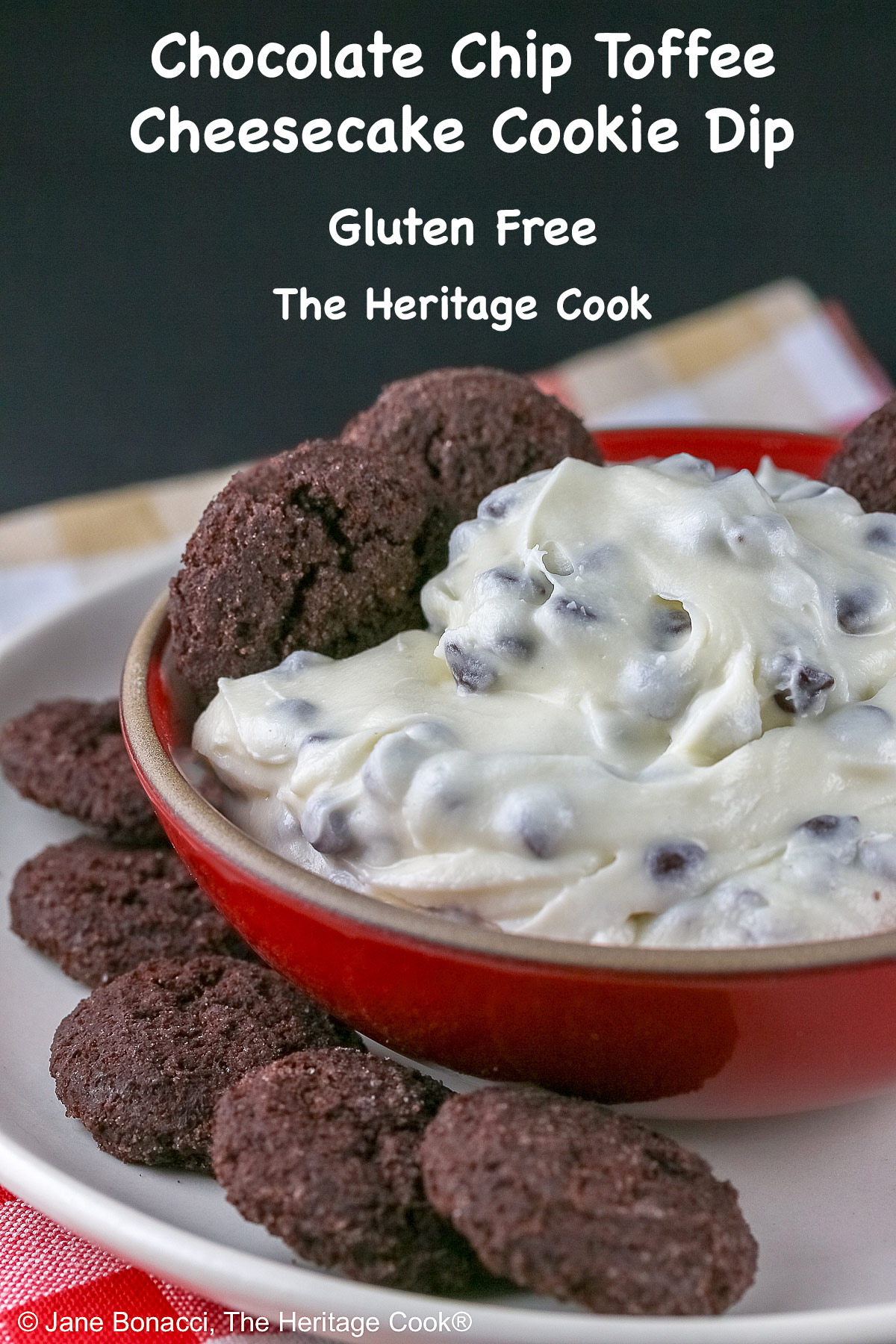 Chocolate Chip Toffee Cheesecake Cookie Dip is piled high in a red bowl on a white plate and surrounded with small chocolate cookies and red-skinned apple slices, all sitting on a red, white, and gold cloth © 2026 Jane Bonacci, The Heritage Cook.