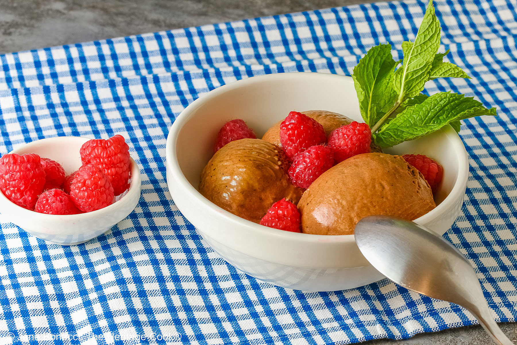 Three scoops of Double Dark Chocolate No-Churn Ice Cream topped with fresh red raspberries and a sprig of mint, sitting on a blue and white checked cloth on a mottled gray background with a spoon resting on the edge of the bowl © 2026 Jane Bonacci, The Heritage Cook.