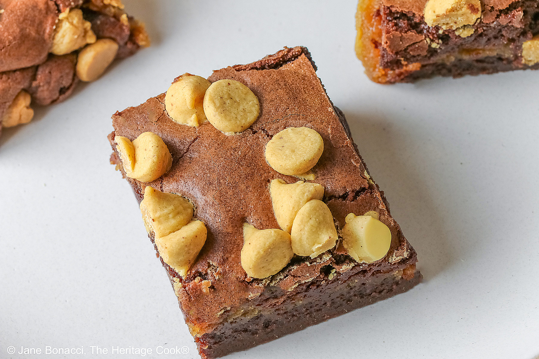 Four Peanut Butter Brownies with a peanut butter layer in the middle and studded with peanut butter chips on top, placed on a white plate with a natural light brown clay border, with denim blue cloths behind, sitting on a neutral gray backdrop © 2026 Jane Bonacci, The Heritage Cook.