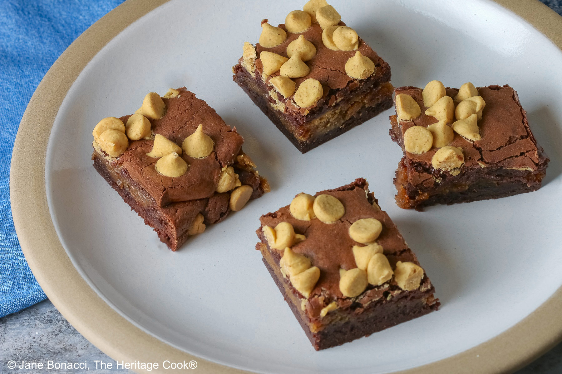 Four Peanut Butter Brownies with a peanut butter layer in the middle and studded with peanut butter chips on top, placed on a white plate with a natural light brown clay border, with denim blue cloths behind, sitting on a neutral gray backdrop © 2026 Jane Bonacci, The Heritage Cook.