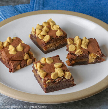 Four Peanut Butter Brownies with a peanut butter layer in the middle and studded with peanut butter chips on top, placed on a white plate with a natural light brown clay border, with denim blue cloths behind, sitting on a neutral gray backdrop © 2026 Jane Bonacci, The Heritage Cook.