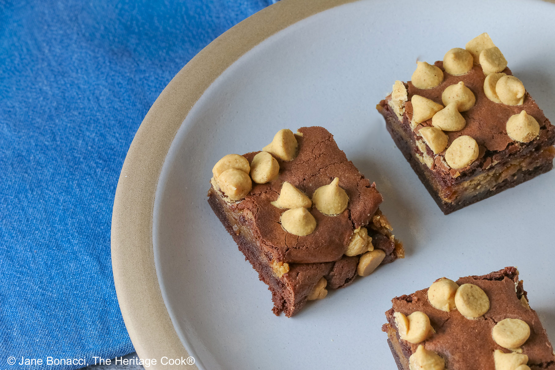 Four Peanut Butter Brownies with a peanut butter layer in the middle and studded with peanut butter chips on top, placed on a white plate with a natural light brown clay border, with denim blue cloths behind, sitting on a neutral gray backdrop © 2026 Jane Bonacci, The Heritage Cook.