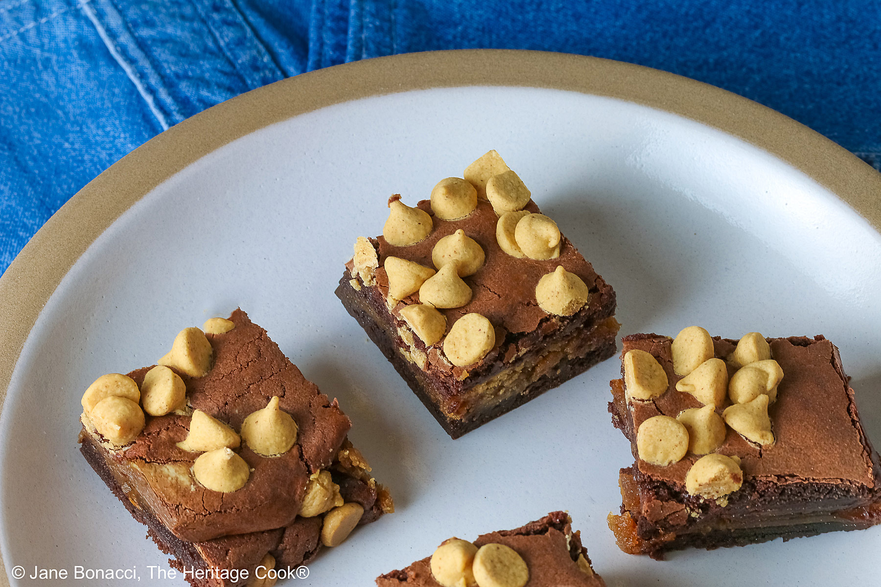 Four Peanut Butter Brownies with a peanut butter layer in the middle and studded with peanut butter chips on top, placed on a white plate with a natural light brown clay border, with denim blue cloths behind, sitting on a neutral gray backdrop © 2026 Jane Bonacci, The Heritage Cook.