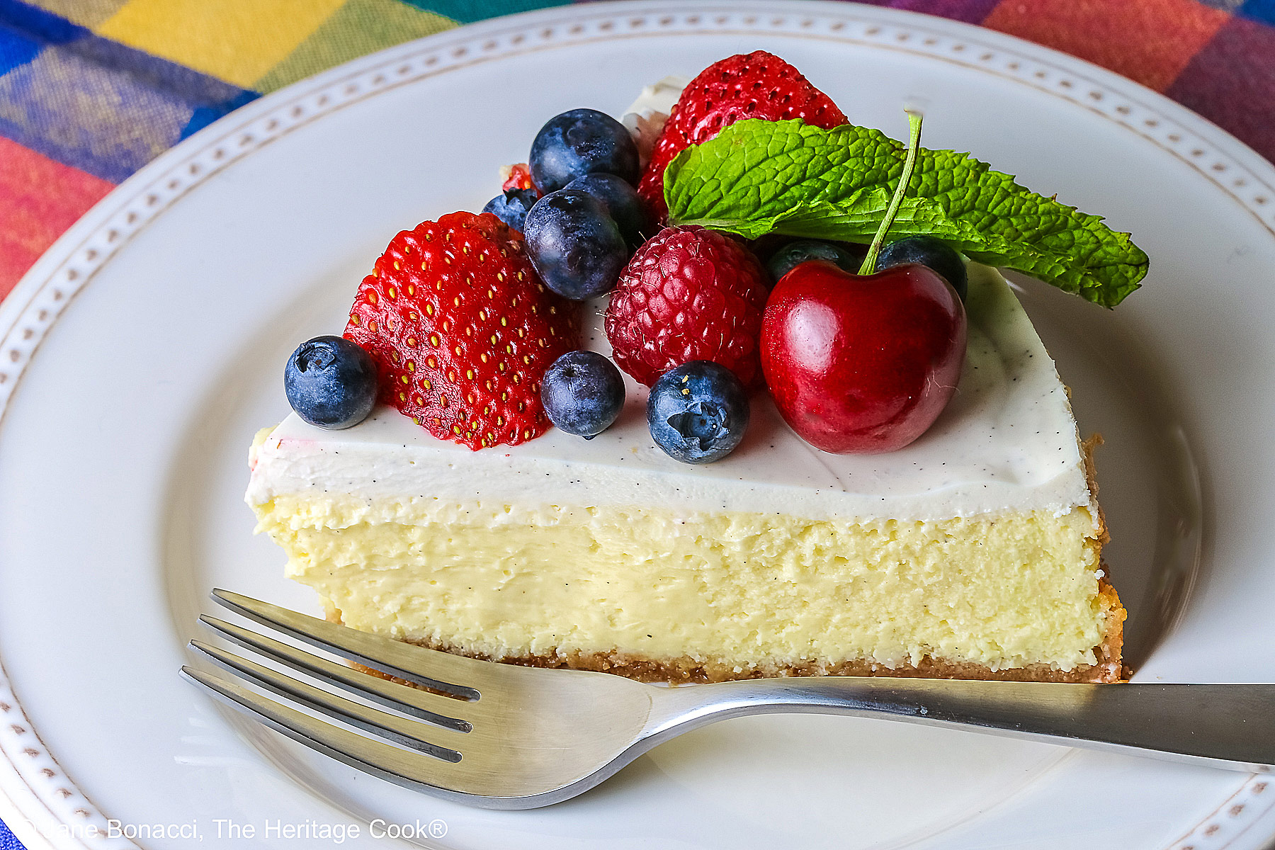 This Red, White, and Blue New York Cheesecake is served with half the top piled high with red and blue fruits, strawberries, raspberries, blueberries, and cherries with a slice cut out and sitting on a creamy white plate which has a brightly multi-colored cloth beneath, some shots with the slice in front and the whole cheesecake behind © 2026 Jane Bonacci, The Heritage Cook.