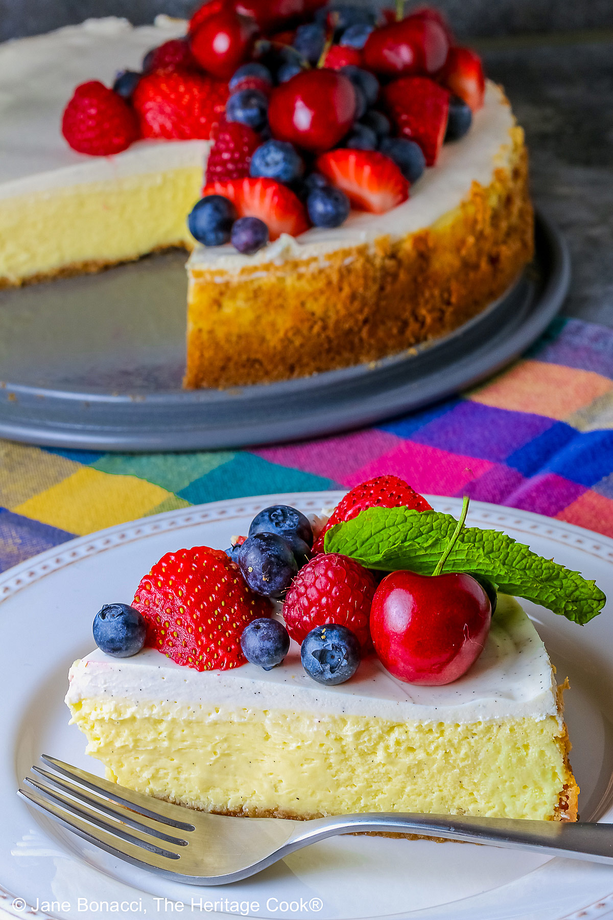 This Red, White, and Blue New York Cheesecake is served with half the top piled high with red and blue fruits, strawberries, raspberries, blueberries, and cherries with a slice cut out and sitting on a creamy white plate which has a brightly multi-colored cloth beneath, some shots with the slice in front and the whole cheesecake behind © 2026 Jane Bonacci, The Heritage Cook.
