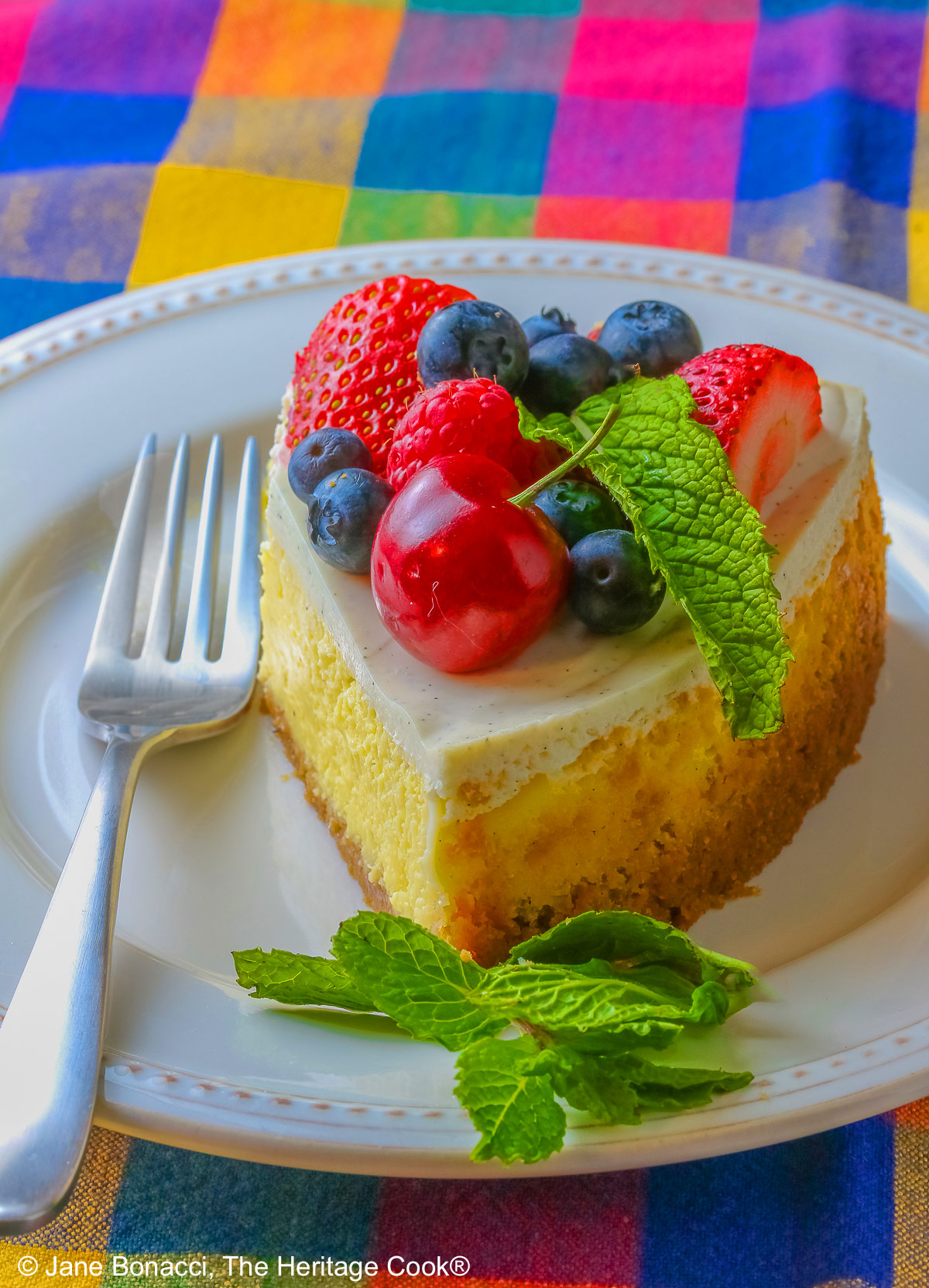 This Red, White, and Blue New York Cheesecake is served with half the top piled high with red and blue fruits, strawberries, raspberries, blueberries, and cherries with a slice cut out and sitting on a creamy white plate which has a brightly multi-colored cloth beneath, some shots with the slice in front and the whole cheesecake behind © 2026 Jane Bonacci, The Heritage Cook.