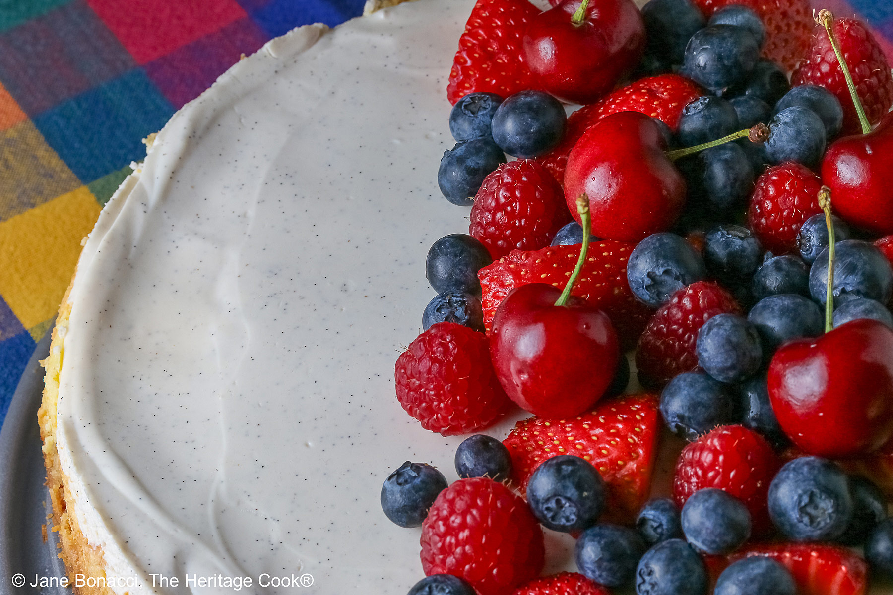 This Red, White, and Blue New York Cheesecake is served with half the top piled high with red and blue fruits, strawberries, raspberries, blueberries, and cherries with a slice cut out and sitting on a creamy white plate which has a brightly multi-colored cloth beneath, some shots with the slice in front and the whole cheesecake behind © 2026 Jane Bonacci, The Heritage Cook.