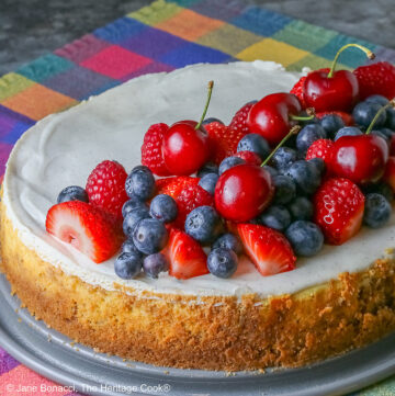 This Red, White, and Blue New York Cheesecake is served with half the top piled high with red and blue fruits, strawberries, raspberries, blueberries, and cherries with a slice cut out and sitting on a creamy white plate which has a brightly multi-colored cloth beneath, some shots with the slice in front and the whole cheesecake behind © 2026 Jane Bonacci, The Heritage Cook.