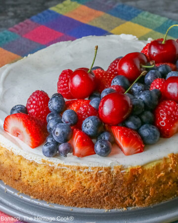 This Red, White, and Blue New York Cheesecake is served with half the top piled high with red and blue fruits, strawberries, raspberries, blueberries, and cherries with a slice cut out and sitting on a creamy white plate which has a brightly multi-colored cloth beneath, some shots with the slice in front and the whole cheesecake behind © 2026 Jane Bonacci, The Heritage Cook.