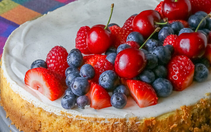 This Red, White, and Blue New York Cheesecake is served with half the top piled high with red and blue fruits, strawberries, raspberries, blueberries, and cherries with a slice cut out and sitting on a creamy white plate which has a brightly multi-colored cloth beneath, some shots with the slice in front and the whole cheesecake behind © 2026 Jane Bonacci, The Heritage Cook.