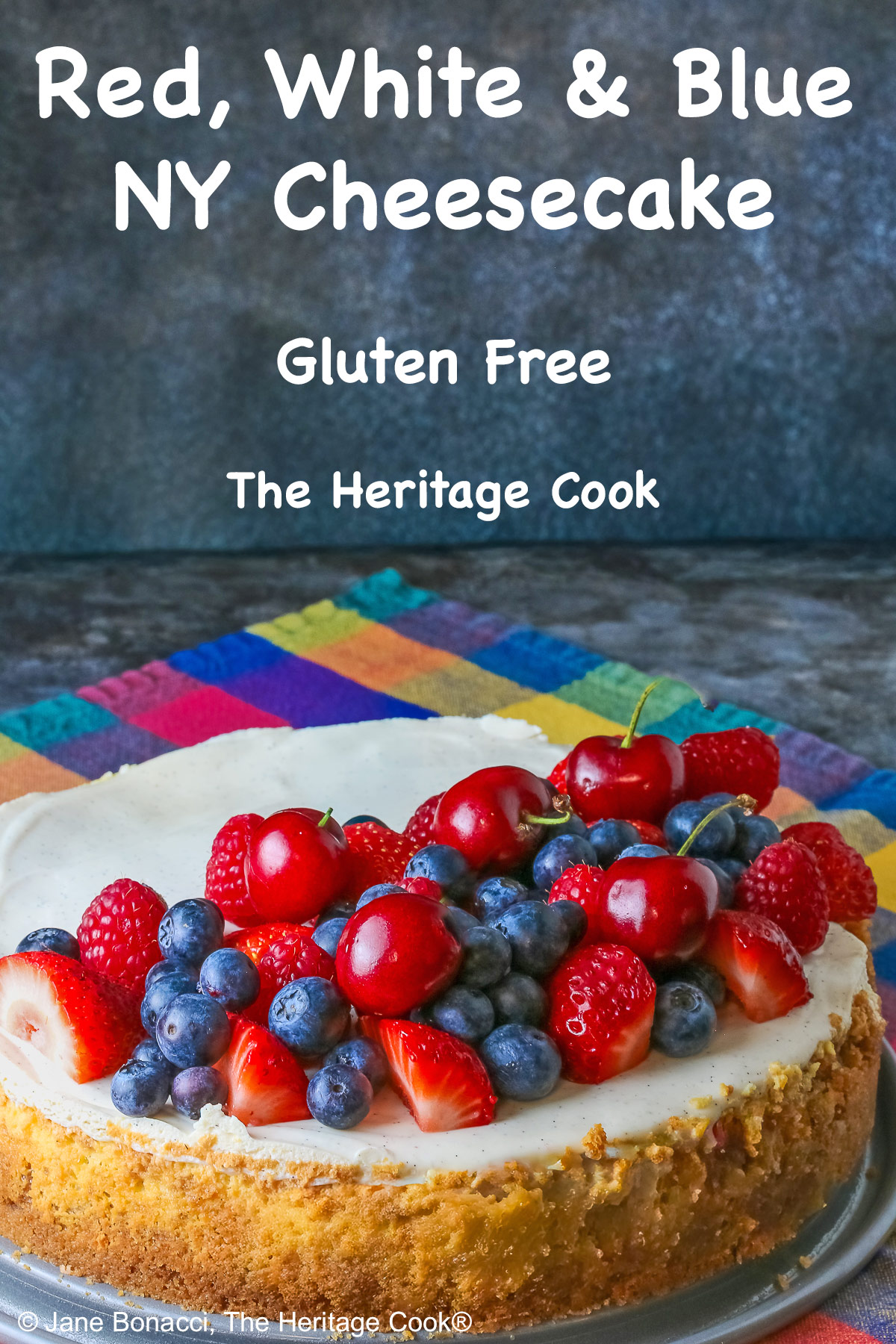 This Red, White, and Blue New York Cheesecake is served with half the top piled high with red and blue fruits, strawberries, raspberries, blueberries, and cherries with a slice cut out and sitting on a creamy white plate which has a brightly multi-colored cloth beneath, some shots with the slice in front and the whole cheesecake behind © 2026 Jane Bonacci, The Heritage Cook.