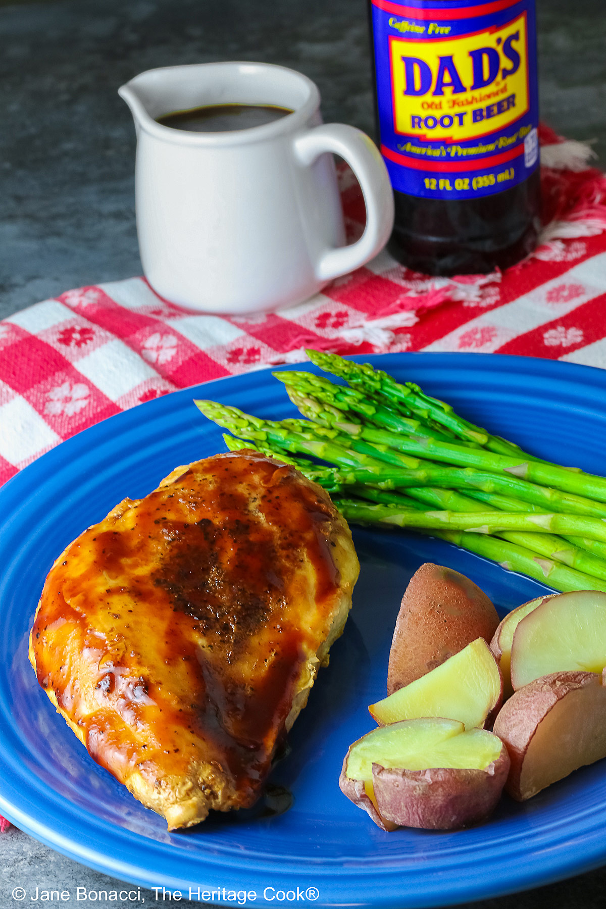 A chicken breast coated with Root Beer BBQ Sauce on a blue plate with asparagus and red potatoes with a small white pitcher of the BBQ sauce and a bottle of root beer behind the plate sitting on a red and white checked cloth © 2026 Jane Bonacci, The Heritage Cook.