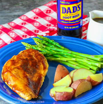 A chicken breast coated with Root Beer BBQ Sauce on a blue plate with asparagus and red potatoes with a small white pitcher of the BBQ sauce and a bottle of root beer behind the plate sitting on a red and white checked cloth © 2026 Jane Bonacci, The Heritage Cook.