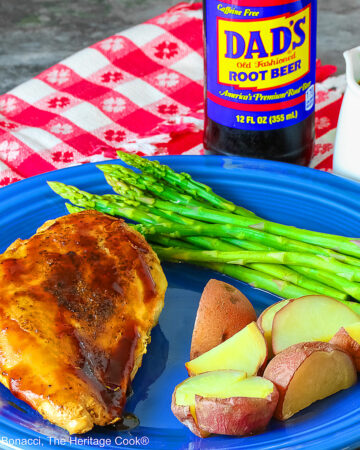 A chicken breast coated with Root Beer BBQ Sauce on a blue plate with asparagus and red potatoes with a small white pitcher of the BBQ sauce and a bottle of root beer behind the plate sitting on a red and white checked cloth © 2026 Jane Bonacci, The Heritage Cook.