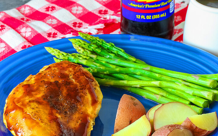 A chicken breast coated with Root Beer BBQ Sauce on a blue plate with asparagus and red potatoes with a small white pitcher of the BBQ sauce and a bottle of root beer behind the plate sitting on a red and white checked cloth © 2026 Jane Bonacci, The Heritage Cook.