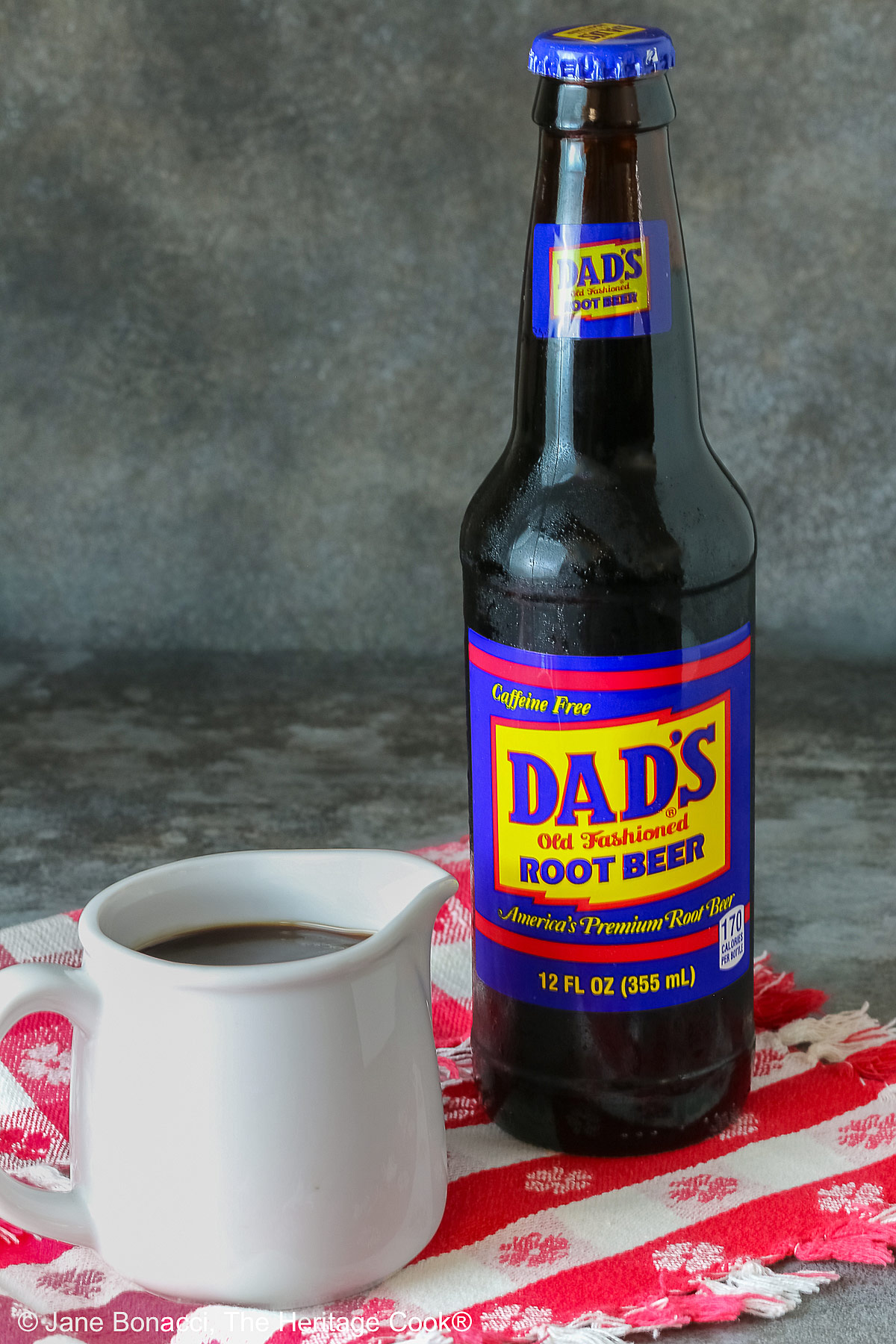 Bottle of root beer next to a small pitcher of the root beer BBQ sauce on a red checked cloth.