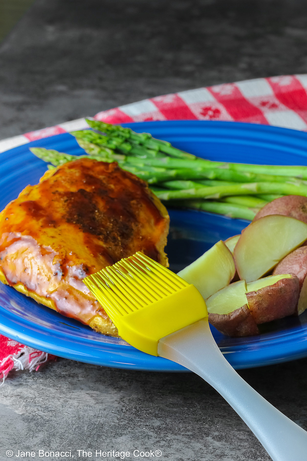 A chicken breast coated with Root Beer BBQ Sauce on a blue plate with asparagus and red potatoes with a yellow pastry brush laying on the chicken after brushing the sauce on top © 2026 Jane Bonacci, The Heritage Cook.