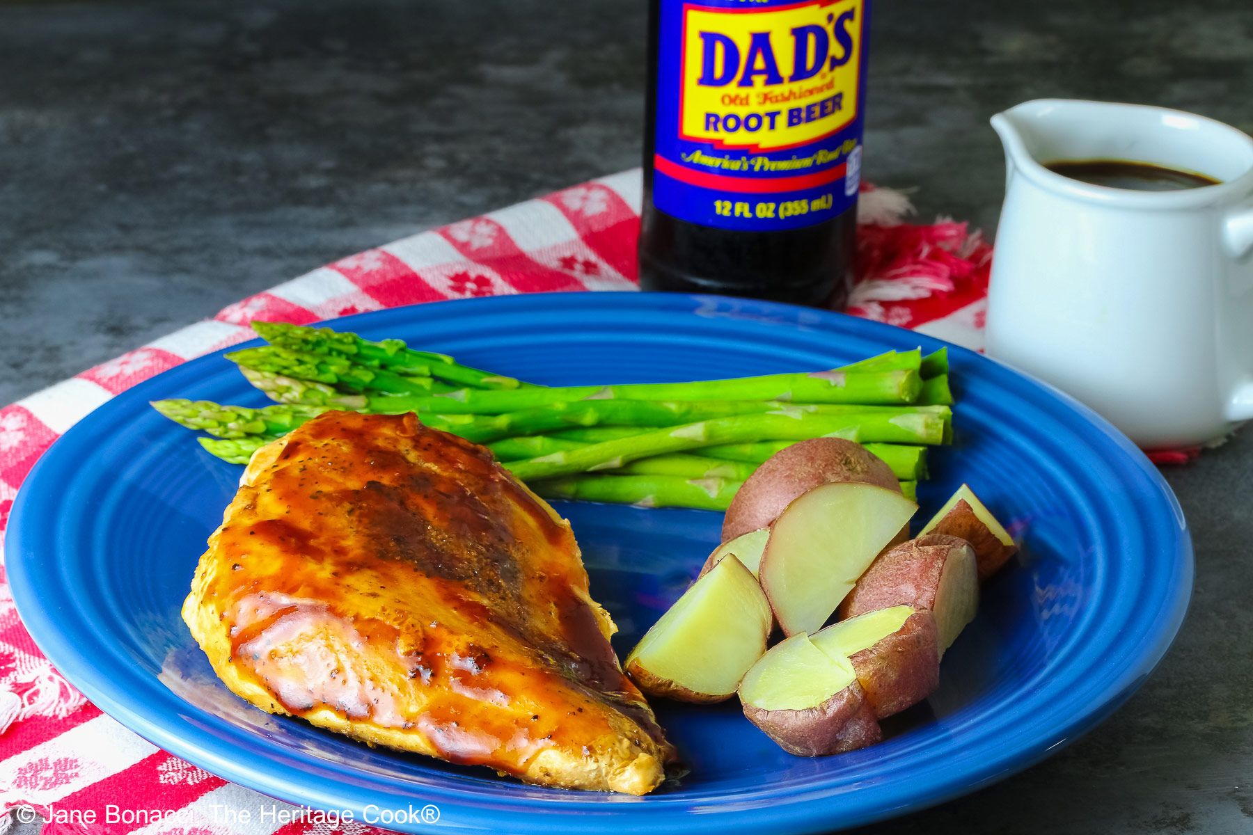 A chicken breast coated with Root Beer BBQ Sauce on a blue plate with asparagus and red potatoes with a small white pitcher of the BBQ sauce and a bottle of root beer behind the plate sitting on a red and white checked cloth © 2026 Jane Bonacci, The Heritage Cook.