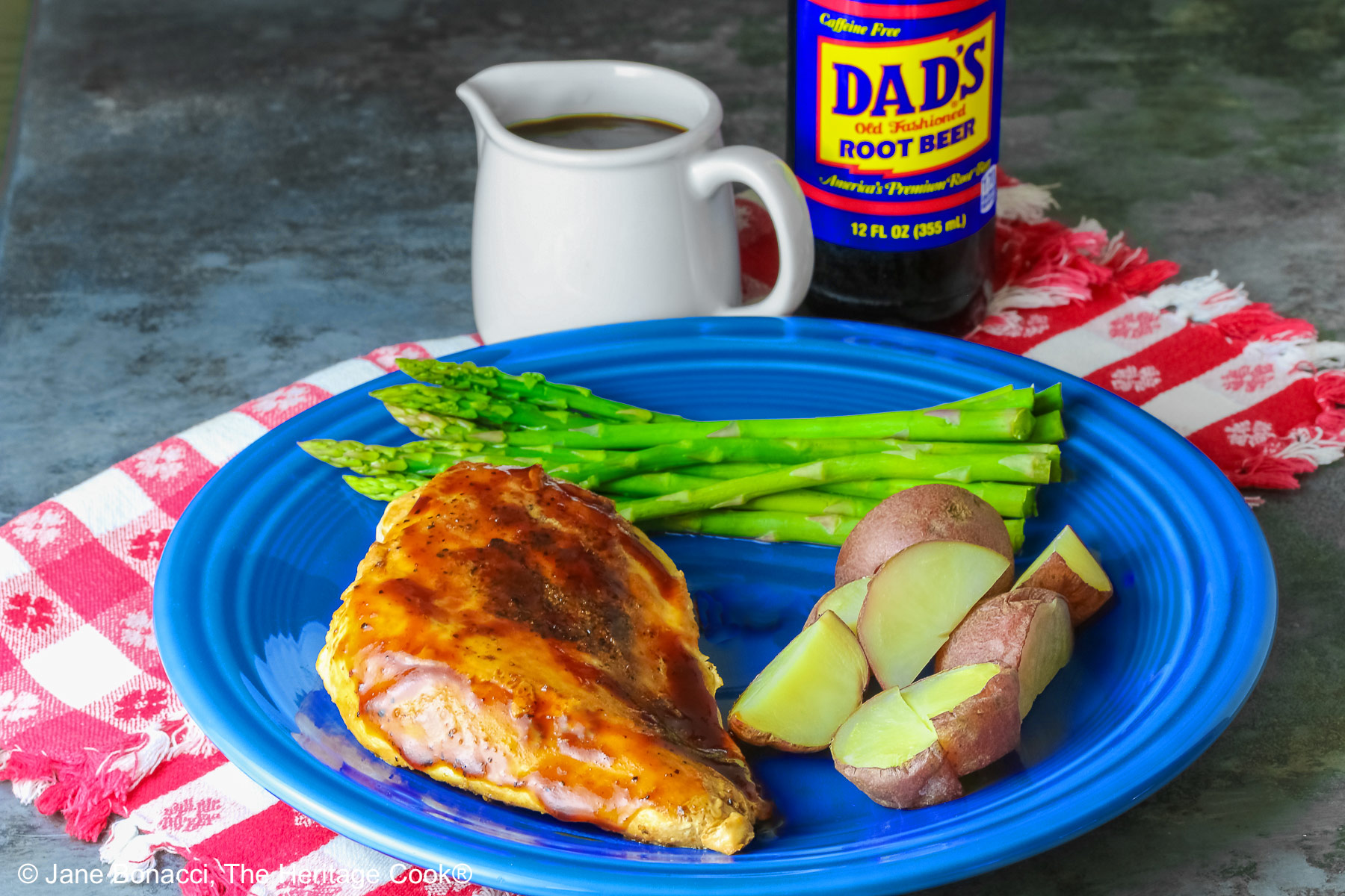 A chicken breast coated with Root Beer BBQ Sauce on a blue plate with asparagus and red potatoes with a small white pitcher of the BBQ sauce and a bottle of root beer behind the plate sitting on a red and white checked cloth © 2026 Jane Bonacci, The Heritage Cook.