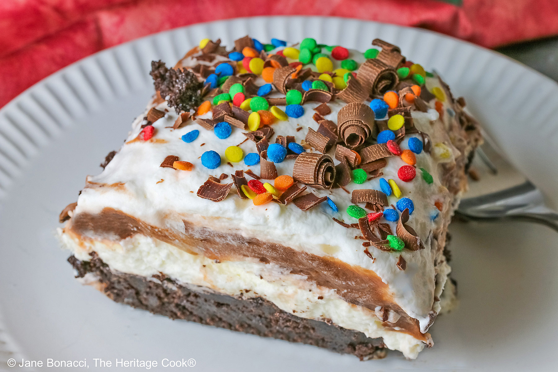 A large square slice of the Brownie Refrigerator Pudding Cake with shaved chocolate and rainbow sprinkles on the top on a white plate with a pink cloth behind it sitting on a marbled gray surface © 2026 Jane Bonacci, The Heritage Cook.