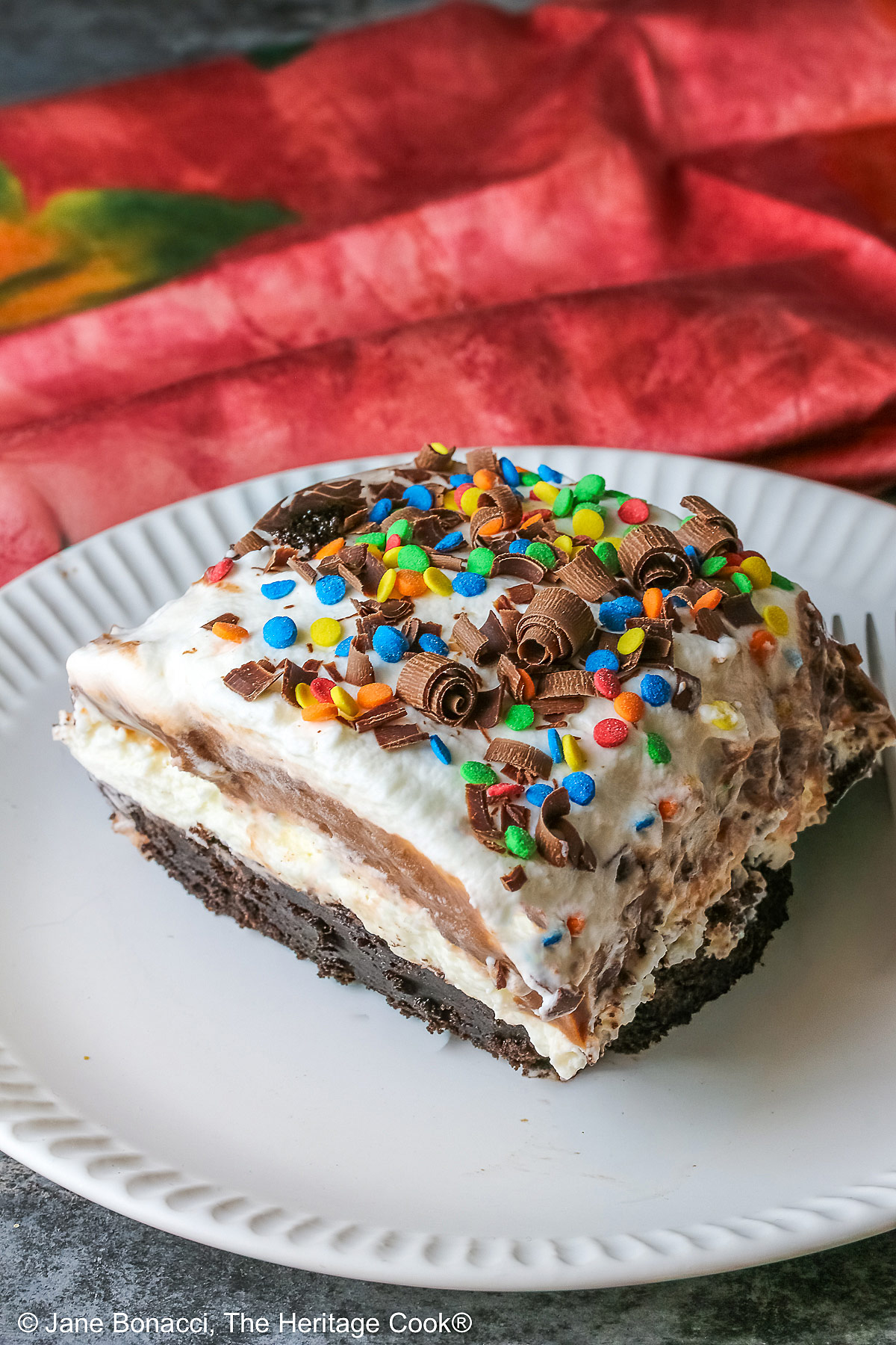 A large square slice of the Brownie Refrigerator Pudding Cake with shaved chocolate and rainbow sprinkles on the top on a white plate with a pink cloth behind it sitting on a marbled gray surface © 2026 Jane Bonacci, The Heritage Cook.