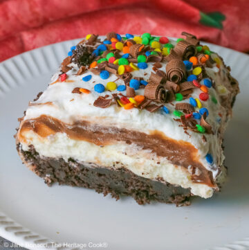 A large square slice of the Brownie Refrigerator Pudding Cake with shaved chocolate and rainbow sprinkles on the top on a white plate with a pink cloth behind it sitting on a marbled gray surface © 2026 Jane Bonacci, The Heritage Cook.